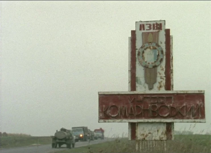 From The East (1993) movie still: wide — A tall, rusted metal monument stands beside a gray paved road under heavy clouds. A line of military trucks…