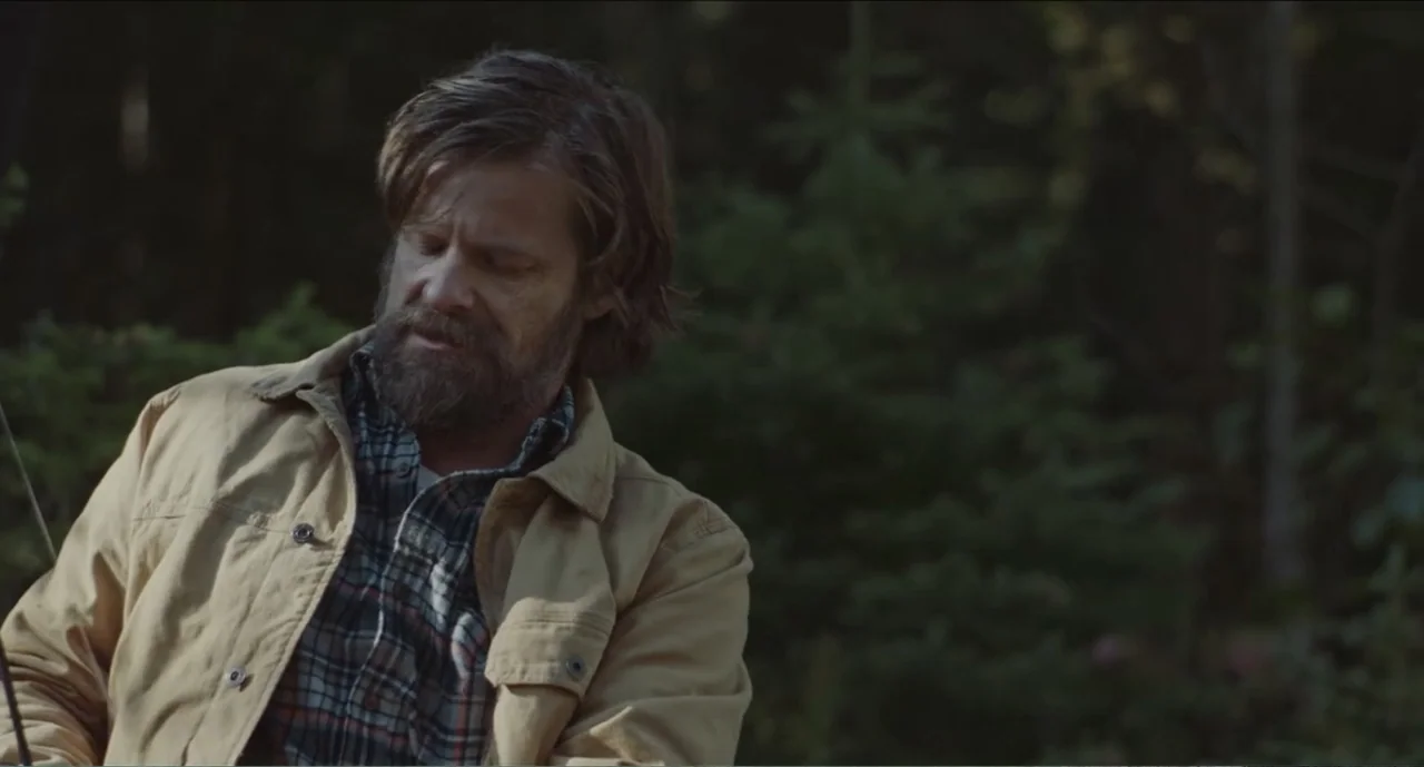 Cowboys (2014) movie still: medium — A man stands outdoors among dark pine trees and foliage. He is looking downwards with a neutral expression…