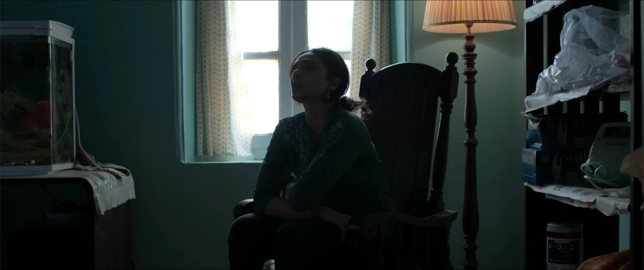 Cheekatilo (2026) movie still: medium — A dimly lit room features a woman sitting in a wooden rocking chair by a window. A tall floor lamp stands…
