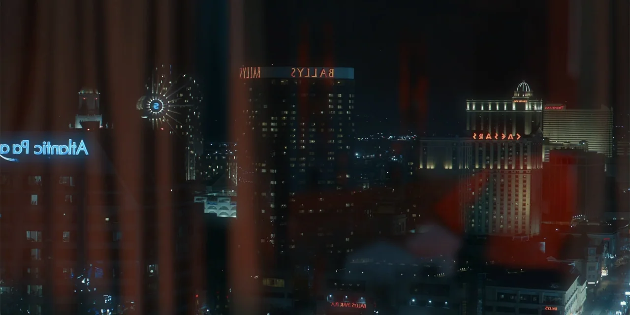 The Pickup (2025) movie still: wide — A nighttime view of a city skyline with glowing neon hotel signs visible through curtains. Multiple high-rise…