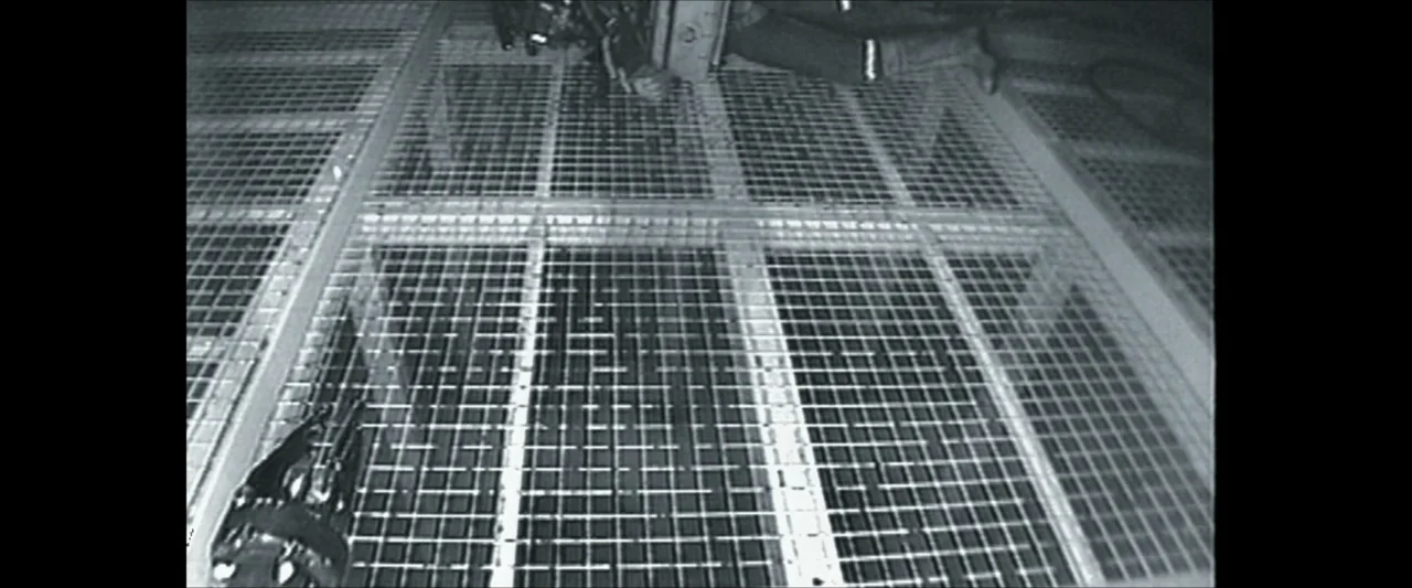 Last Breath (2025) movie still: wide — A metal grating walkway is illuminated by overhead industrial lights in a dark space. The floor consists of…