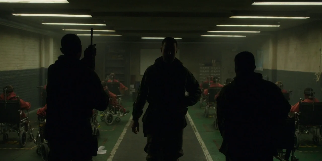 The Girl With All The Gifts (2016) movie still: fullbody — A dimly lit industrial basement contains rows of stationary individuals sitting in metal…