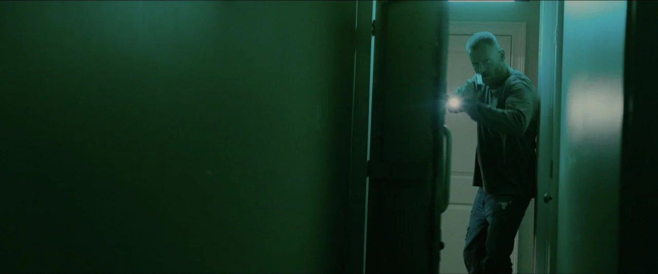Ambush (2020) movie still: medium — A dimly lit indoor hallway is shrouded in a heavy green-tinted color grade. A man holding a flashlight peeks…