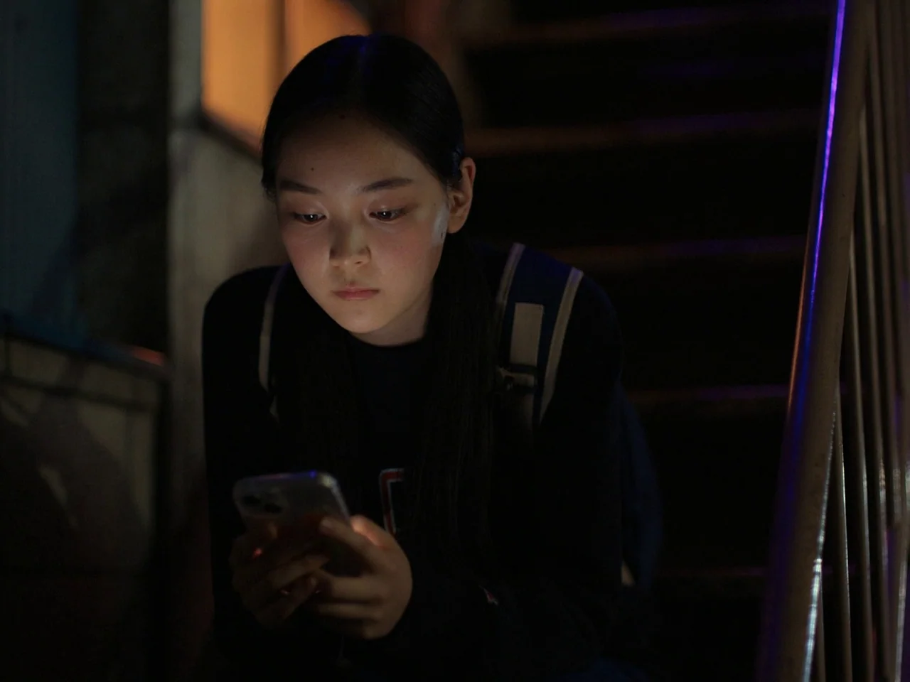 Perfect Days (2023) movie still: medium — A dimly lit indoor stairwell at night with wooden steps visible in the background. A young woman with a…