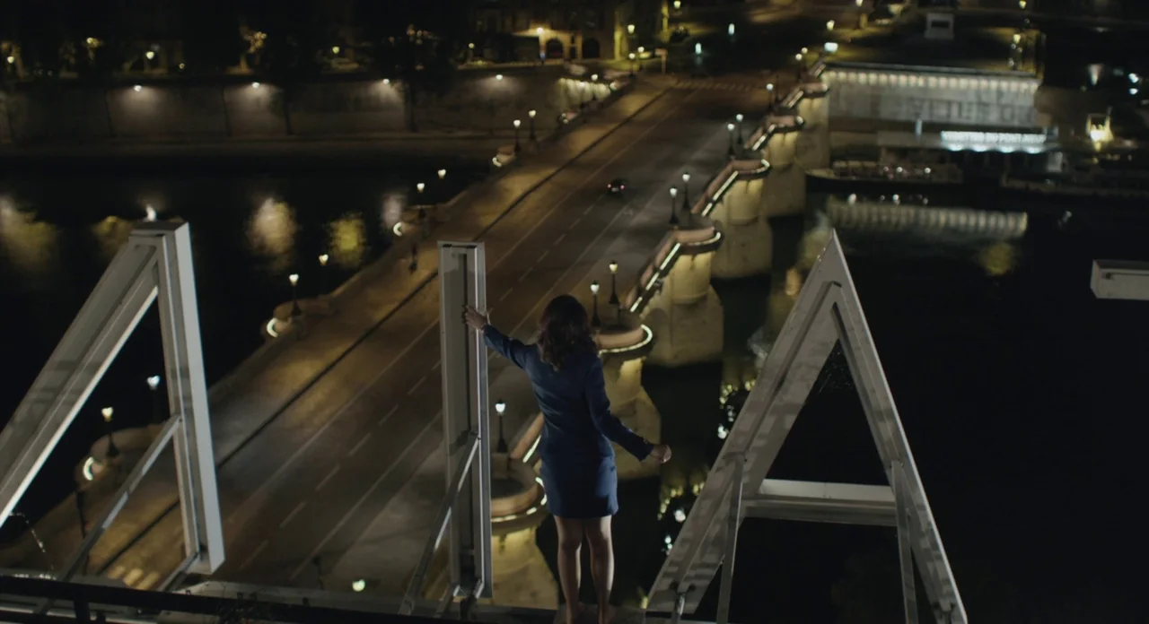 Holy Motors (2012) movie still: wide — A woman stands on the metal frame of a large rooftop sign at night. Below her, a bridge spans a dark river…