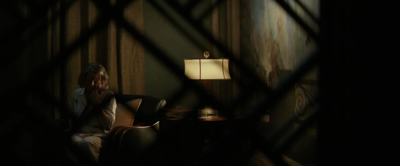 Hotel Artemis (2018) movie still: medium — A dimly lit interior space features a woman sitting in a dark leather armchair. A table lamp with a…