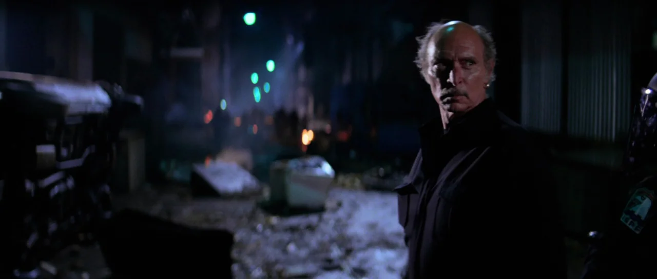 Escape From New York (1981) movie still: medium — A dark, littered urban space sits in the background under artificial night lighting. An older man…