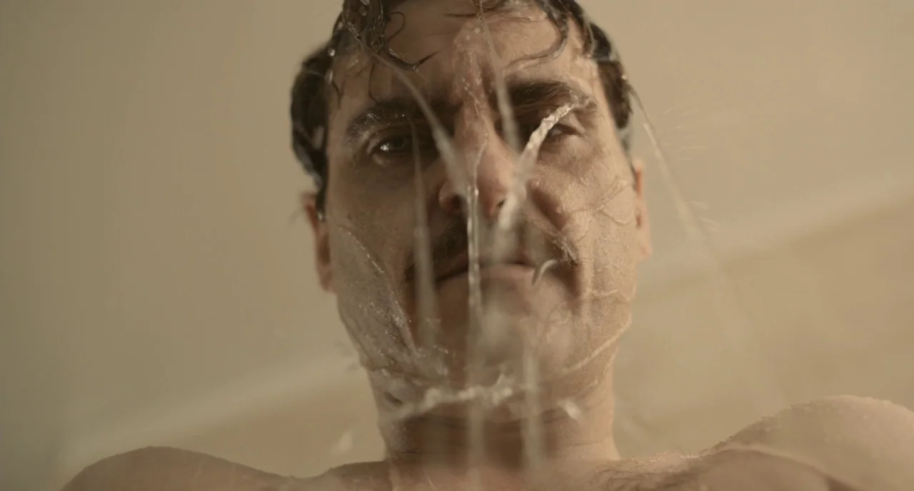 Her (2013) movie still: closeup — A man stands in a brightly lit shower with pale tiled walls. Water runs directly over his face and hair.