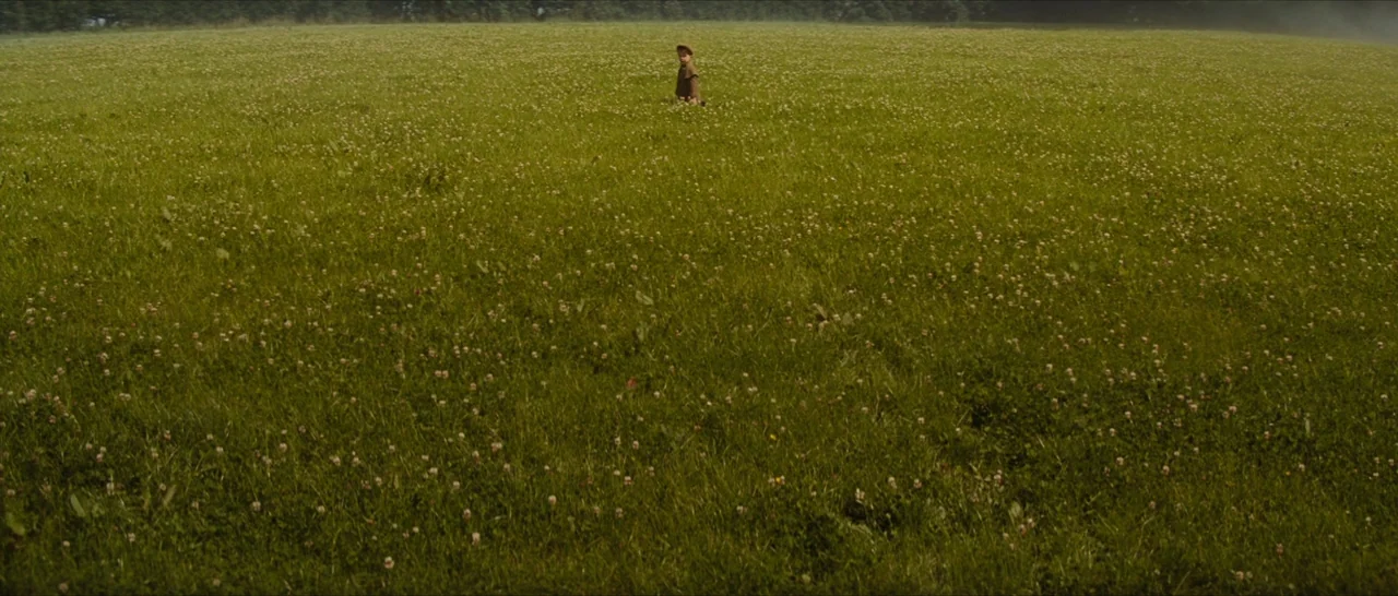 The Lost City of Z (2016) movie still: wide — A vast, flat grassy field stretches toward a dense treeline in the distance. A single young child…