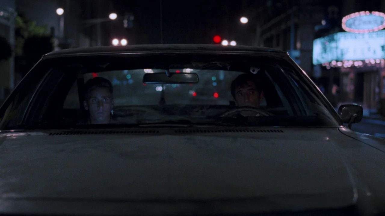 Repo Man (1984) movie still: medium — Two men sit in the front seats of a vehicle at night. The city street lights and a glowing neon sign are…