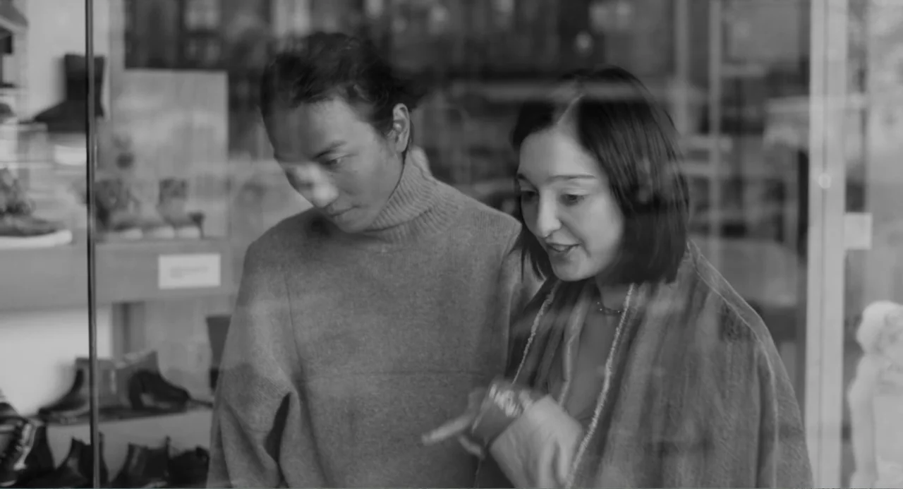 El Planeta (2021) movie still: medium — A sidewalk in front of a retail shop window displays several shoes behind glass. A man and woman stand…