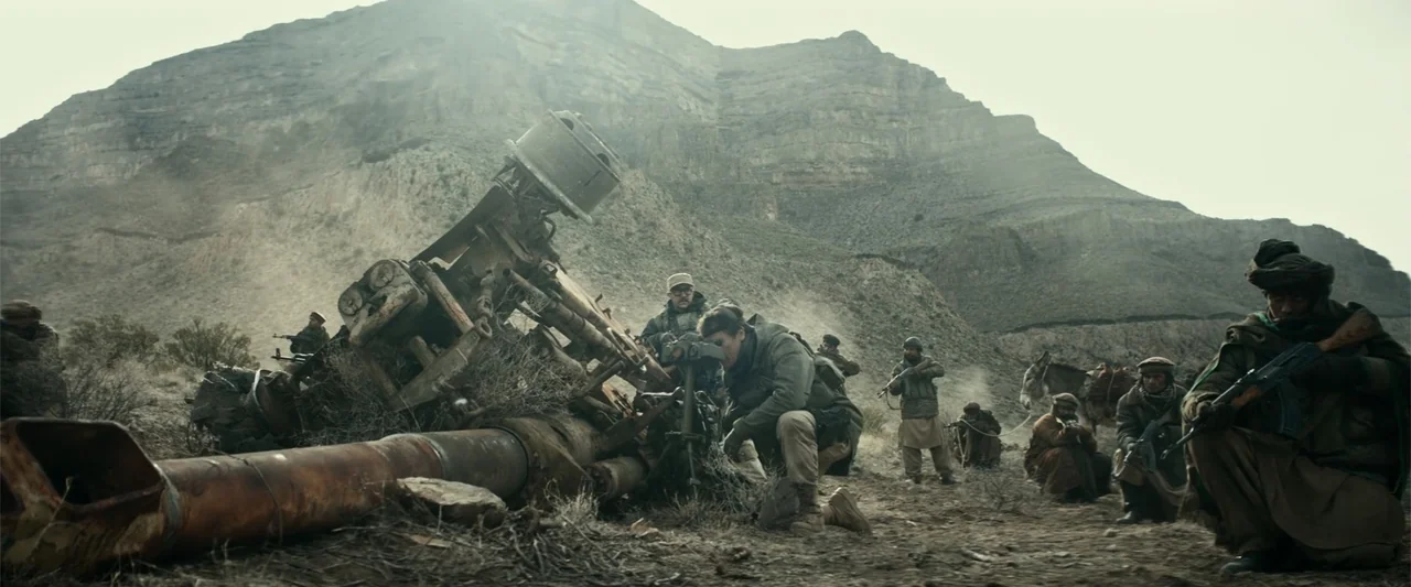 12 Strong (2018) movie still: wide — A rugged mountain landscape features a large damaged artillery piece in the foreground. Several soldiers and…