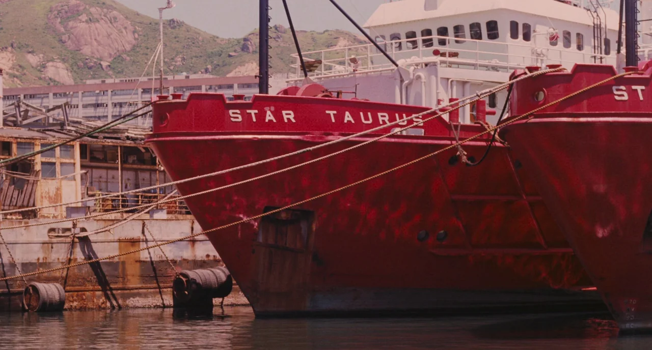 A Better Tomorrow (1986) movie still: medium — A large red industrial boat is tied to a concrete pier in a harbor. Hills and industrial buildings…