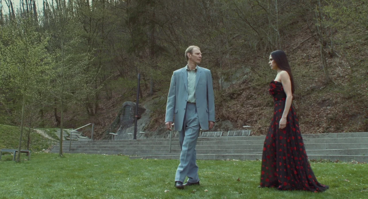 Pina (2011) movie still: fullbody — A man and woman stand on a green lawn in front of concrete steps. A forest with leafless trees rises on the…