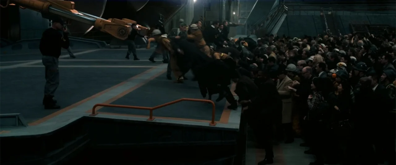 2012 (2009) movie still: wide — A large industrial concrete loading dock at night with a crowd of people. A dense group of people presses against a…