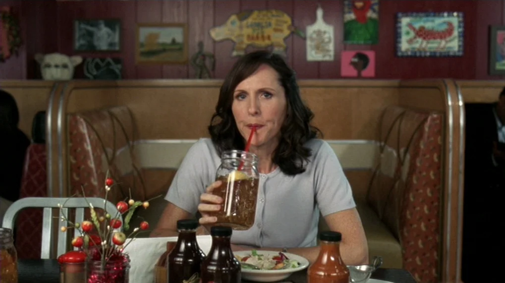 Year Of The Dog (2007) movie still: medium — A woman sits in a diner booth with red wood paneled walls decorated with art. She holds a glass mason…