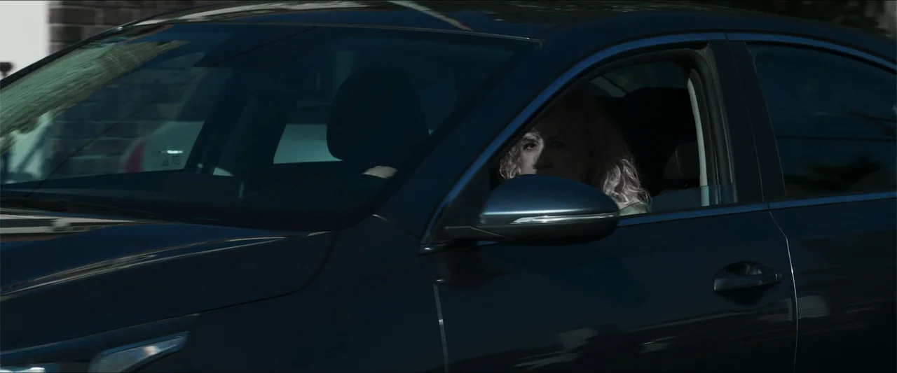 Los habitantes (2023) movie still: medium — A dark sedan is positioned outdoors during daylight with clear reflections on the windows. A woman with…