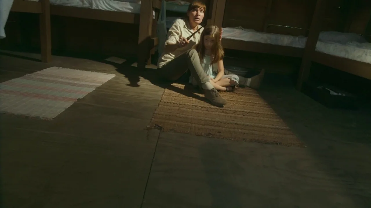 The Sacrament (2013) movie still: medium — A dimly lit room contains wooden bunk beds and woven rugs on the floor. An adult woman sitting on the rug…