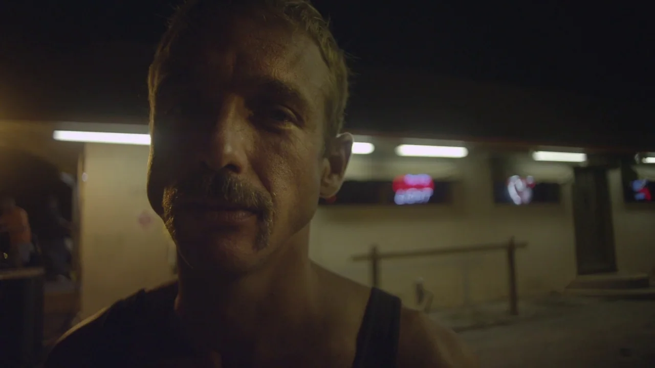 Florida Man (2015) movie still: closeup — A man stands outdoors at night in front of a building with fluorescent lights. He looks directly at the…