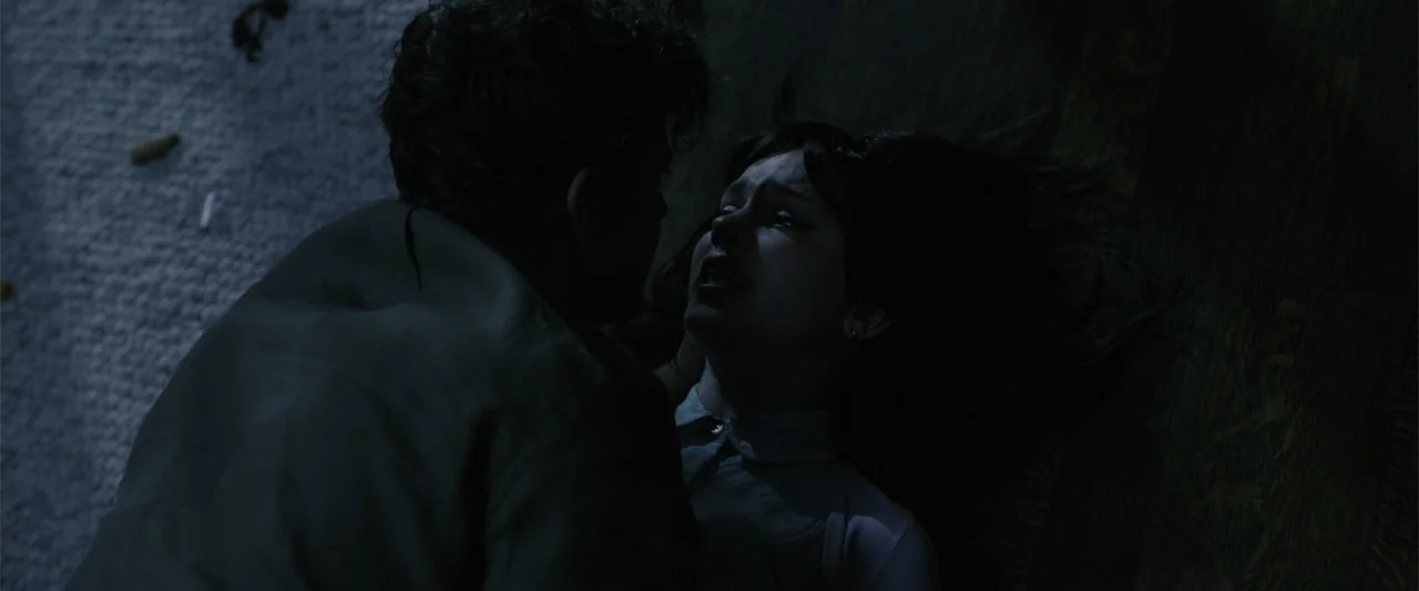 Companion (2025) movie still: medium — A dimly lit interior space with a man and a woman on the floor. The woman reclines while the man leans over…