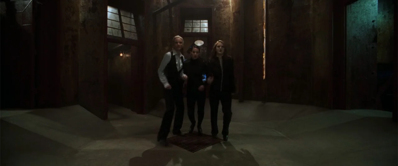 Charlie's Angels (2000) movie still: fullbody — An industrial hallway with weathered, textured metal walls and a dim concrete ground surface. Three…