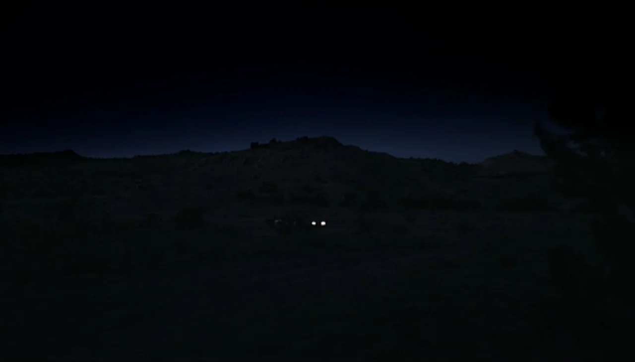 Dark Country (2009) movie still: wide — A vast, dark desert landscape spans the frame under a deep night sky. A pair of vehicle headlights glows…