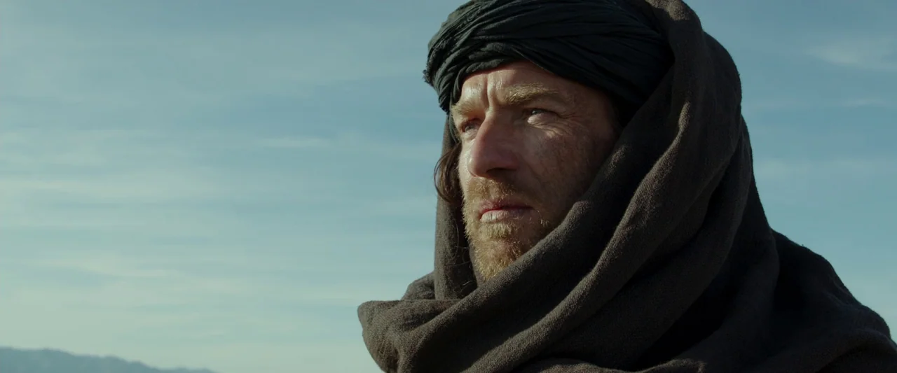 Last Days in the Desert (2015) movie still: closeup — An open desert landscape stretches out under a clear, bright blue sky. A man wears a heavy…