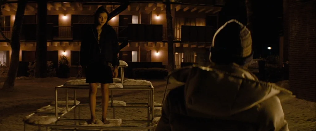 Let Me In (2010) movie still: fullbody — A nighttime view of a two-story motel with warm lights glowing against the facade. A girl in a hoodie…