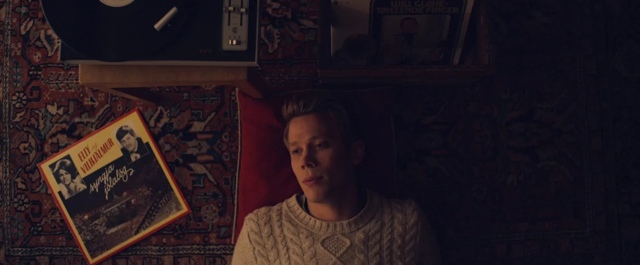 Rift (2017) movie still: medium — A dark, warm-toned room features a vintage vinyl record player and record covers spread out. A young man in a…