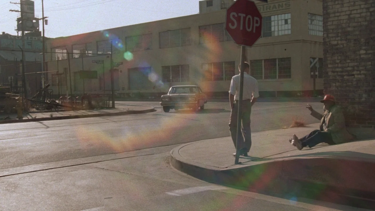 Repo Man (1984) movie still: wide — A quiet urban street corner features a stop sign and an industrial building background. A man stands at the curb…