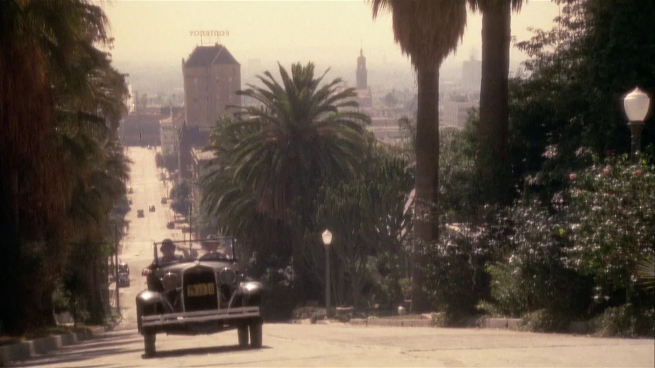 The Day of the Locust (1975) movie still: wide — A vintage convertible drives down a sloping street lined with tall palm trees. In the distance, the…