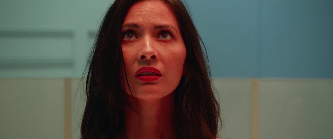 The Predator (2018) movie still: closeup — An interior space features pale cyan walls divided by vertical wooden panels. A woman looks upward while…