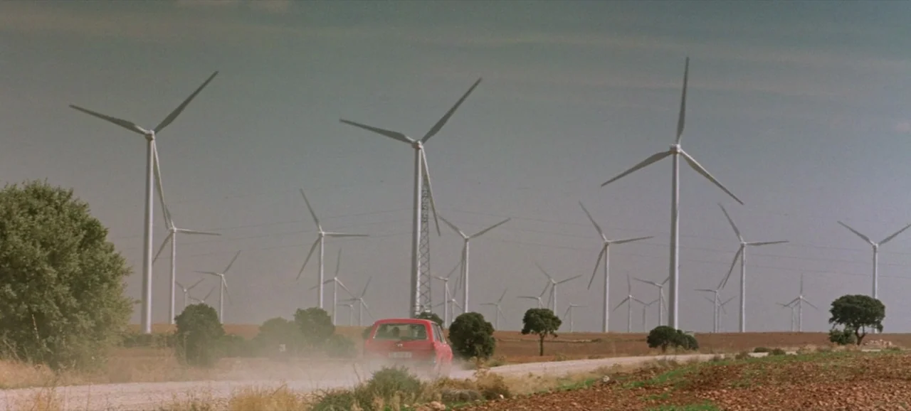 Volver (2006) movie still: wide — A sprawling wind farm with many turbines occupies a vast, dry, open landscape. A small red car moves along a dirt…