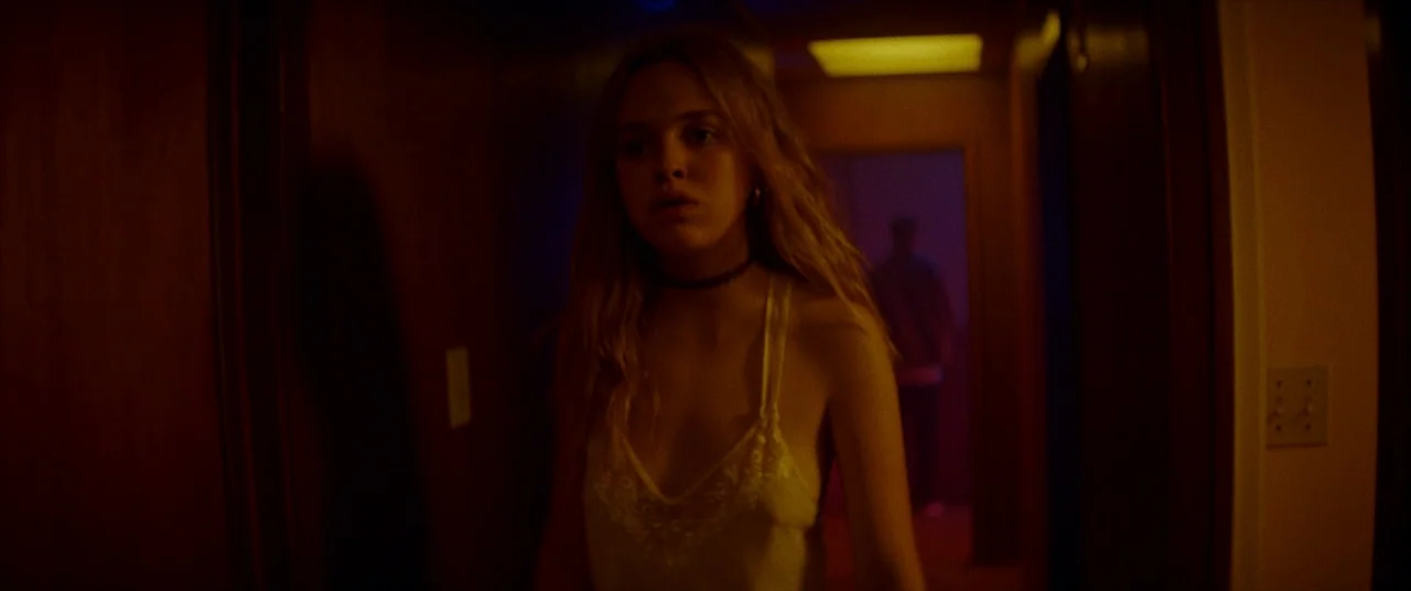 Assassination Nation (2018) movie still: medium — A dim narrow residential hallway features wooden doors and a muted yellow light. A young woman…