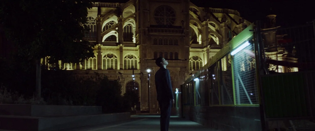 Nocturama (2016) movie still: fullbody — A man stands before a large, floodlit stone cathedral at night. Construction fencing with bright green…