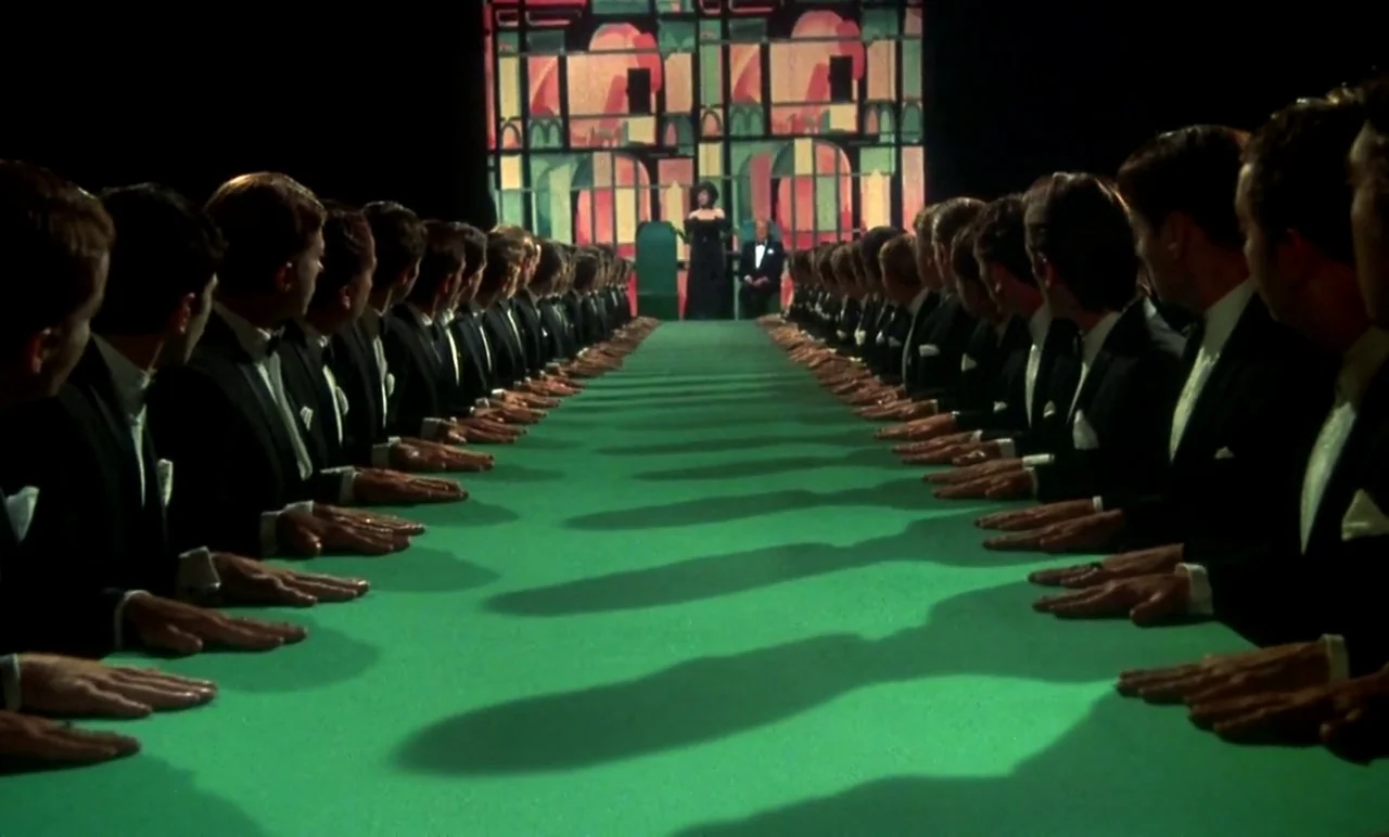 New York, New York (1977) movie still: wide — A long, green-covered table stretches toward a distant stage with a stained-glass backdrop. Two rows…