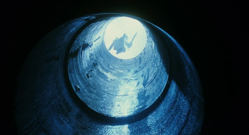 Mimic (1997) movie still: wide — A dark, cylindrical concrete tunnel stretches upward toward a bright, circular opening above. A lone figure is…