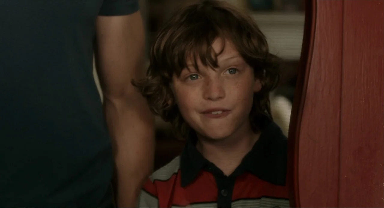Blended (2014) movie still: medium — A domestic interior with a reddish wooden frame visible in the foreground. A young boy stands next to an adult…