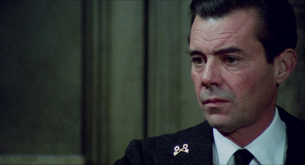 The Night Porter (1974) movie still: closeup — A man stands in an office with dark paneled walls in the background. The man looks toward the side of…