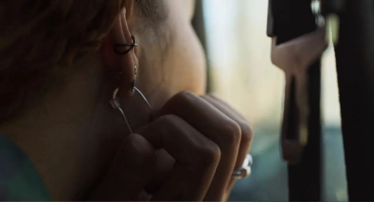 Salvation (2025) movie still: closeup — Interior of a car with sunlight hitting the side of a person's head. A woman adjusts her metal hoop earring…
