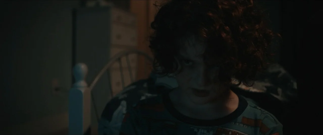 Possessions (2024) movie still: closeup — A dark bedroom interior features a child positioned in front of a metal bed frame. The child stares…