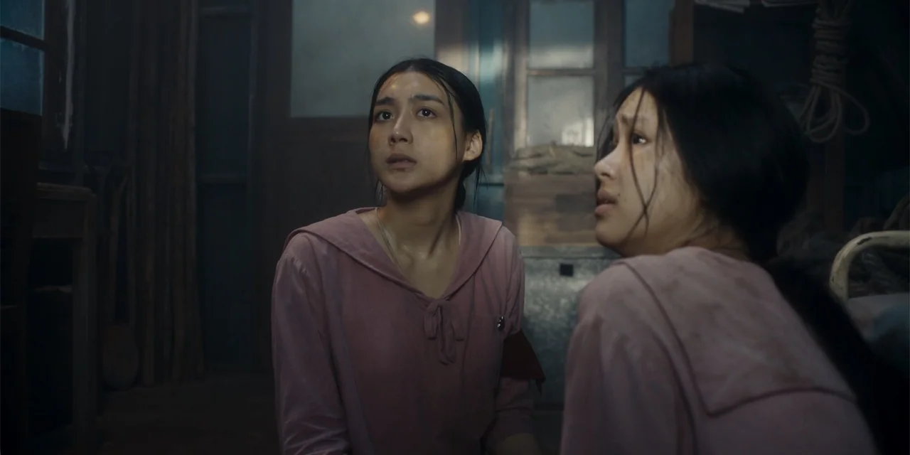 Host (2020) movie still: medium — A dimly lit room features wood paneling and a worn, rustic atmosphere. Two young women in matching pink shirts…
