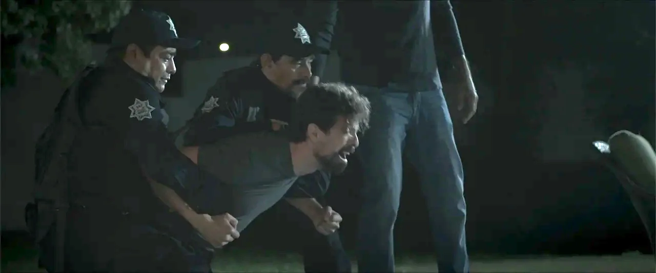 Los habitantes (2023) movie still: medium — A dark night setting with two uniformed officers restraining a shouting man. The men stand in a group as…