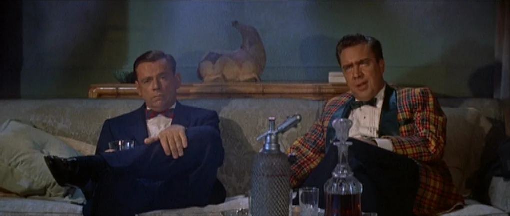 The Girl Can’t Help It (1956) movie still: medium — Two men sit together on a patterned sofa in a dimly lit room. They hold glasses while sitting…