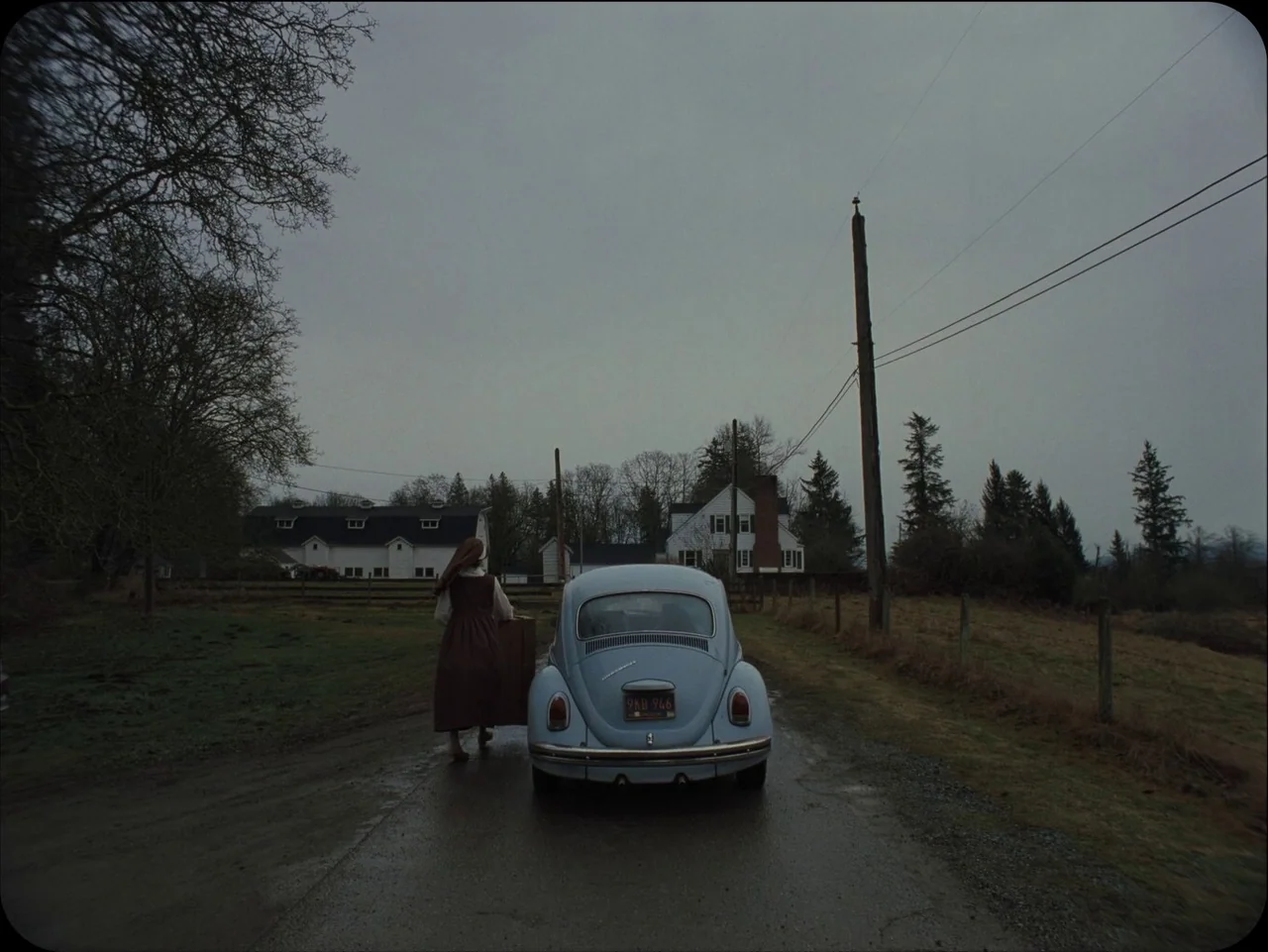 Longlegs (2024) movie still: wide — A rural landscape with a wet asphalt road flanked by fields and houses. A woman in a long brown dress carries a…