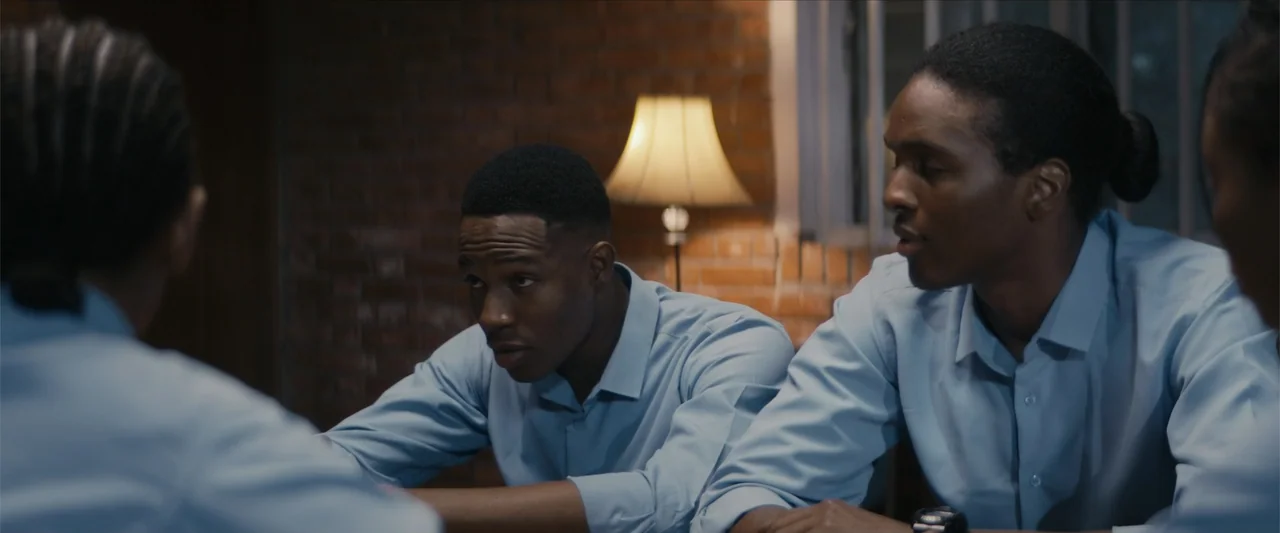Ms. Kanyin (2025) movie still: medium — An interior space with a red brick wall illuminated by a single warm lamp. Three young men in matching light…