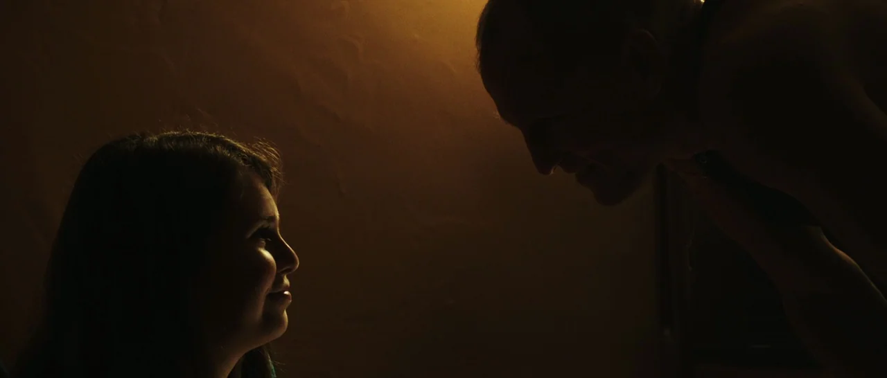 Rampart (2011) movie still: medium — A dimly lit indoor space features dark, warm-toned walls and shadow-filled corners. A younger woman and an…
