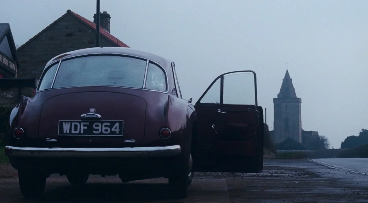 Phantom Thread (2017) movie still: wide — A dark red vintage car sits on the shoulder of a grey paved road. In the distance, a tall stone church…