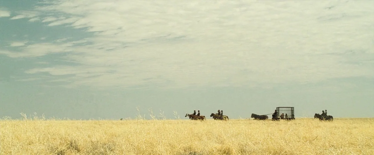 The Proposition (2005) movie still: wide — A vast, flat expanse of dry yellow grass stretches toward a pale sky. A small caravan of riders and a…