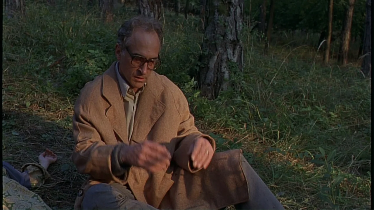 Citizen X (1995) movie still: medium — A dense woodland area with tall trees and overgrown grass under golden light. An older man wearing a tan coat…